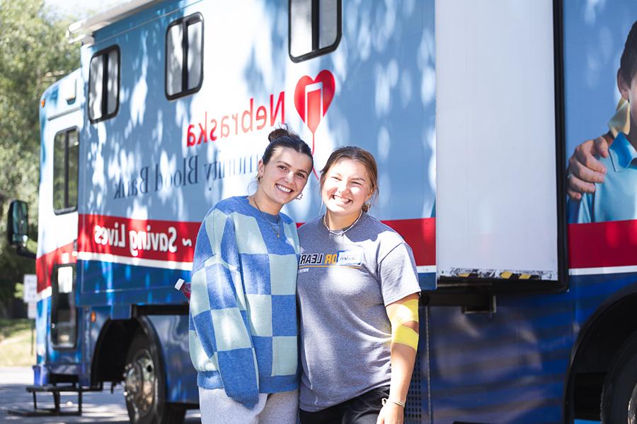 Two students, one with a yellow bandage around her elbow after donating blood, stand in front of the Nebraska Community Blood Bank's Bloodmobile outside of Perry Campus Center in Crete. 